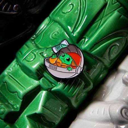 a pin with a green alien in a gray space ship