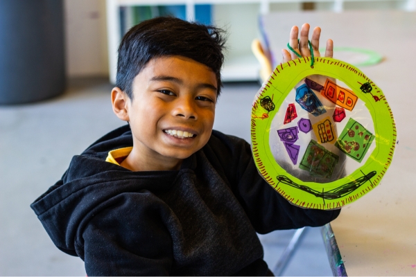 a boy smiles and holds up an art project he made during Kids Art Lab