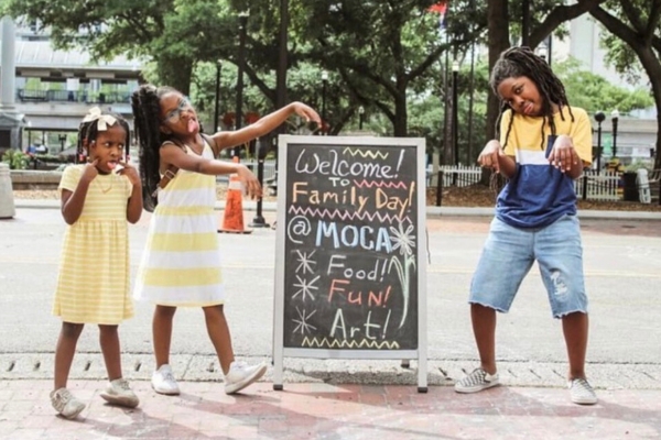 three children make funny faces and point to a chalk board that says it's MOCA Family Day 