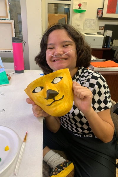 a teen smiling and posing with their artwork, which is a mask that looks like a lion