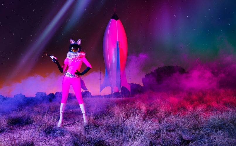 Photograph by Cara Romero of a figure in a bright pink spacesuit standing before a rocket, with pink and purple haze across a desert landscape. click for more information about this exhibition
